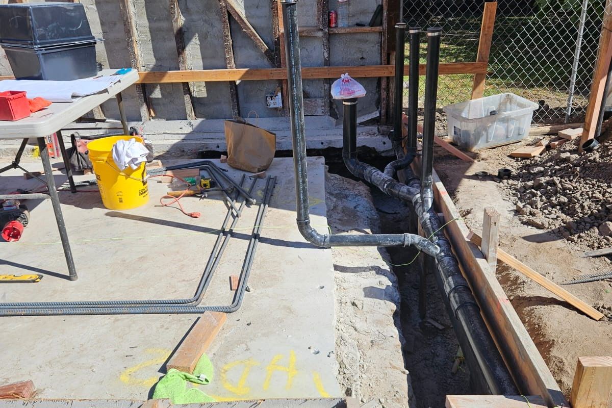 Black drain and vent pipes laid in a trench beside the foundation forms of a backyard unit